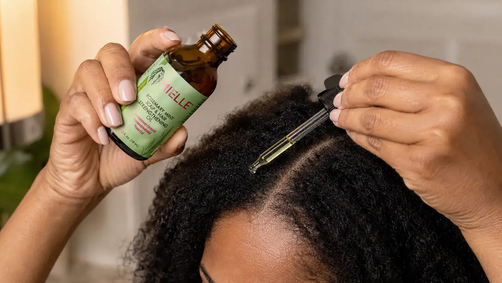 Hands applying a few drops of Mielle Rosemary Mint Scalp Oil to a part in thick natural hair